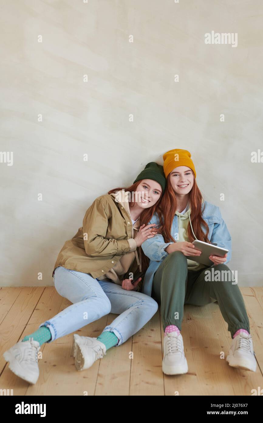 Vertikales, hochwinkeliges Studioporträt von fröhlichen jungen Zwillingen, die stilvolle, lässige Outfits tragen, die zusammen auf Flor sitzen und die Kamera lächeln Stockfoto
