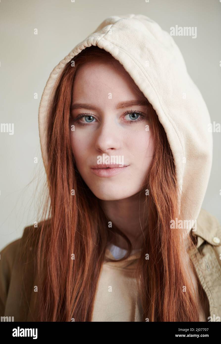 Vertikale Nahaufnahme der attraktiven jungen kaukasischen Frau mit roten Haaren und grauen Augen, die eine Kapuze tragen und die Kamera betrachten Stockfoto
