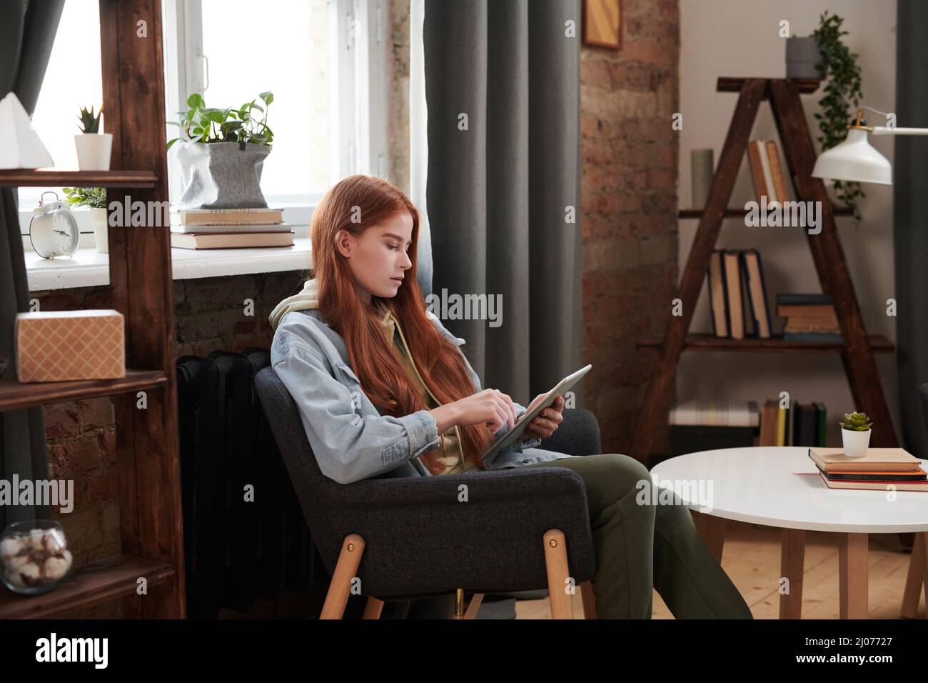Horizontale mittlere lange Seitenansicht Aufnahme der schönen Frau mit langen roten Haaren sitzen entspannt im Stuhl im Wohnzimmer Surfen Interent auf digitalem Tablet Stockfoto