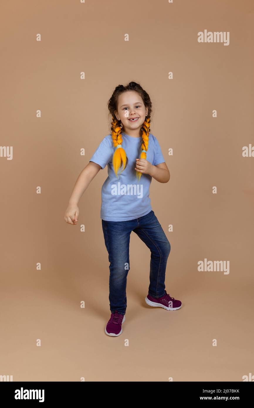 Aufgeregt weibliche Kind Blick auf Kamera lächelnd mit Zähnen tanzen bewegliche Arme mit gelben Kanekalon Zöpfe auf dem Kopf auf beigem Hintergrund tragen blau Stockfoto