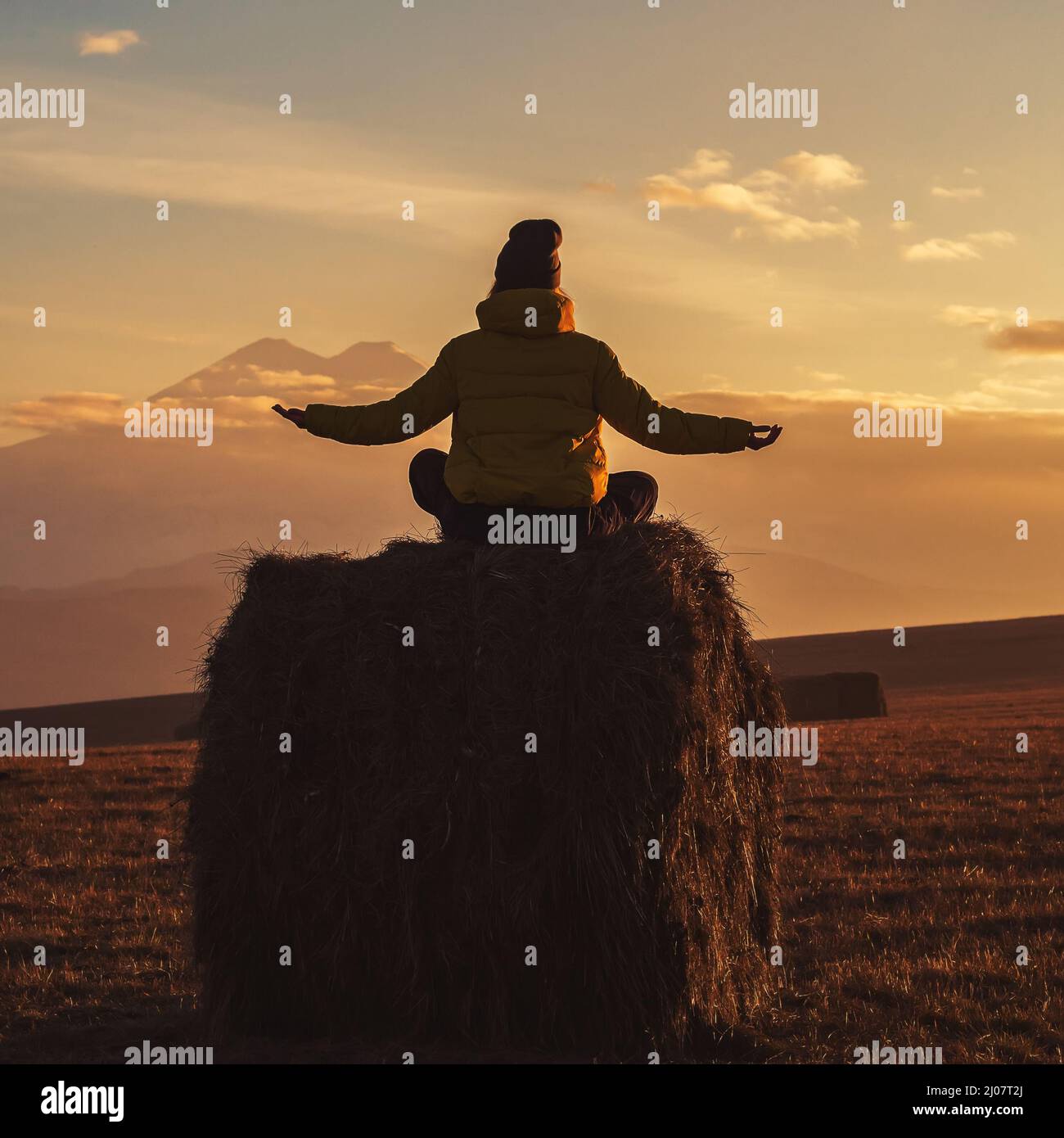 Eine Frau sitzt auf dem Heuhaufen und breitet ihre Handflächen an den Seiten vor dem Hintergrund von Elbrus in den Wolken bei Sonnenuntergang aus. Blick von hinten Stockfoto