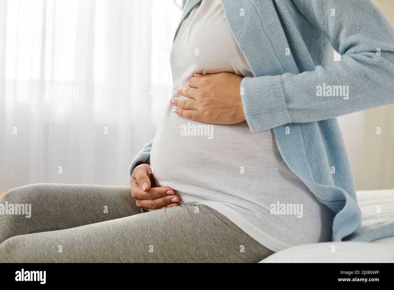 Glückliche junge, schwanger Frau, die auf dem Bett sitzt und ihre Hände auf ihrem Bauch hält Stockfoto