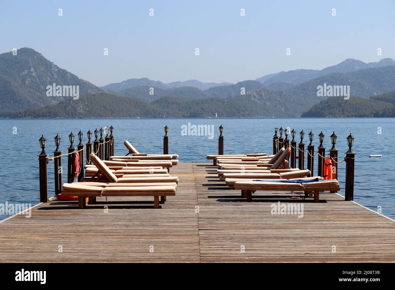 Strandurlaub am Meer Resort in der Ägäis Küste der Türkei, Liegestühle auf einem hölzernen Pier gegen Berge. Leere Sonnenliegen auf grünem Uferhintergrund Stockfoto
