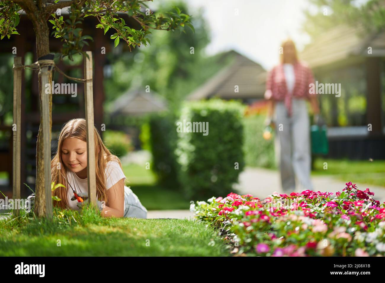 Hübsche weibliche Teenager in Handschuhen, die sich mit ihrer Mutter um Hinterhofpflanzen kümmert. Kaukasische Frau, die auf dem Hintergrund läuft, während ihre Tochter Gartenarbeit leistet. Stockfoto