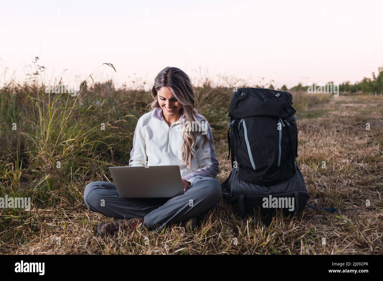 Junge Frau mit einem Rucksack neben ihr, die mitten auf dem Feld einen Laptop für die Telearbeit benutzt. Digitaler Nomade. Stockfoto