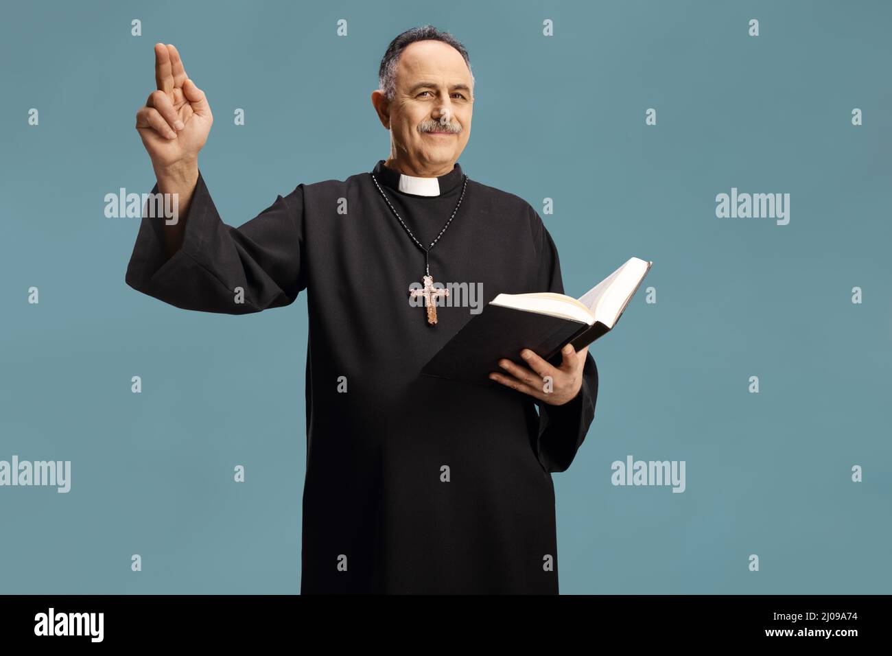 Reifer Priester liest ein Gebet aus der bibel und gestikiert mit Hand isoliert auf blauem Hintergrund Stockfoto