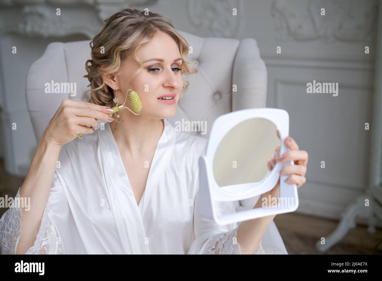 Schöne Frau in Seidenrobe mit Jade Gesichtswalze mit natürlichen Steinen tun Massage halten Spiegel in der Hand, während im Sessel sitzen morgendliche Routine. Hautpflege Schönheitsbehandlung Konzept zu Hause Stockfoto