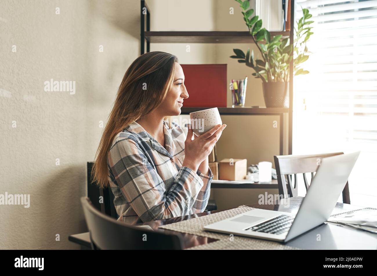 Zuerst Kaffee, dann nicht tun etwas Browsen. Eine kleine Aufnahme einer jungen Frau, die morgens zu Hause Kaffee trinkt und einen Laptop benutzt. Stockfoto