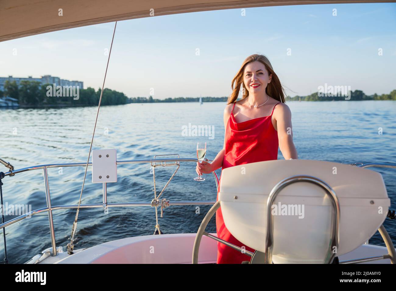 Die pritty Frau in einem langen roten Kleid mit einem Glas Champagner auf einer Yacht stehend und auf der Suche nach Blick auf den Sonnenuntergang am Fluss Stockfoto