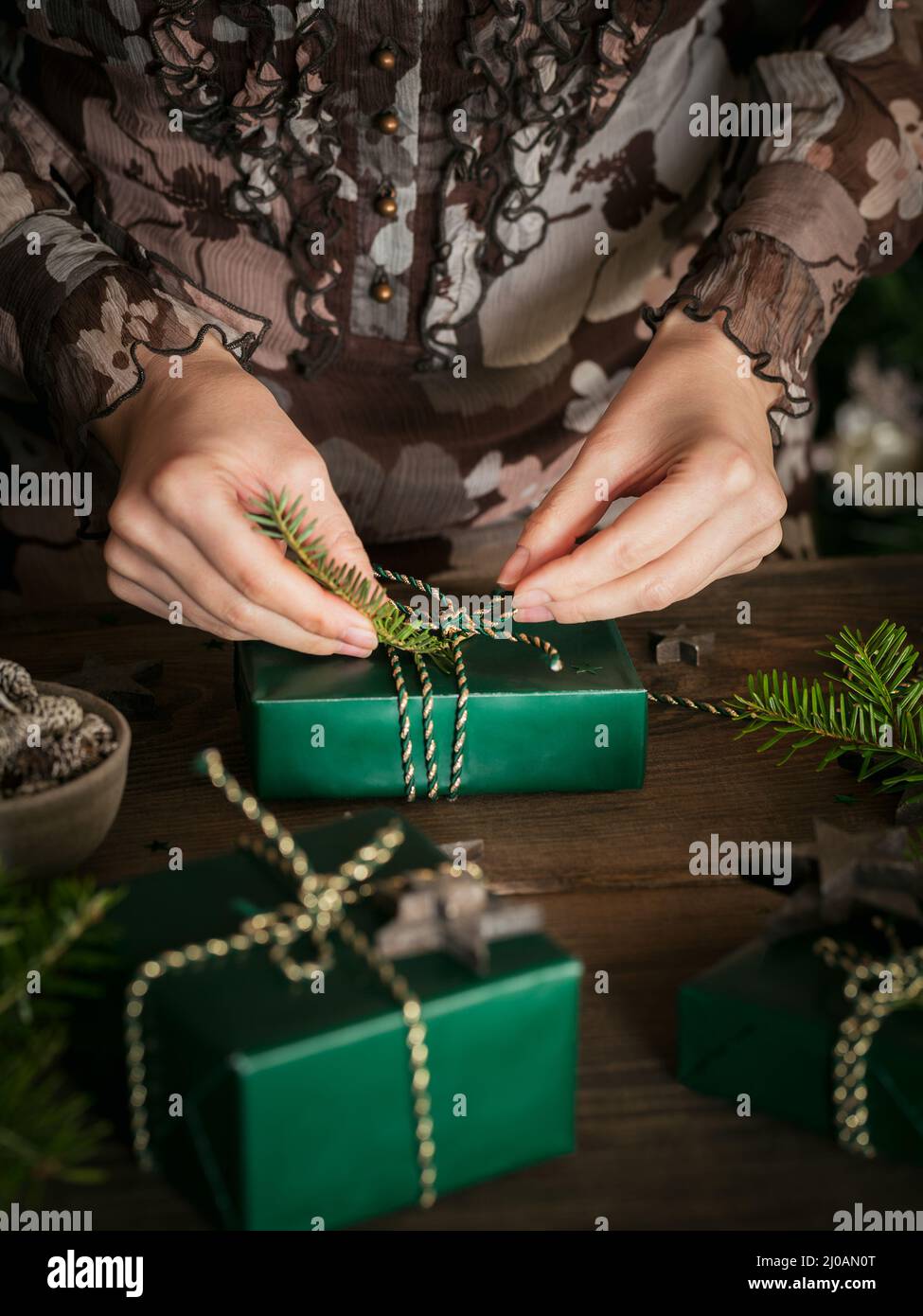 Frau macht schöne Weihnachtsgeschenk am Tisch. Hände halten stilvolle einfache Weihnachtsgeschenk in grünem Papier auf rustikalen dunklen Tisch mit Tannenzweigen, Stift Stockfoto