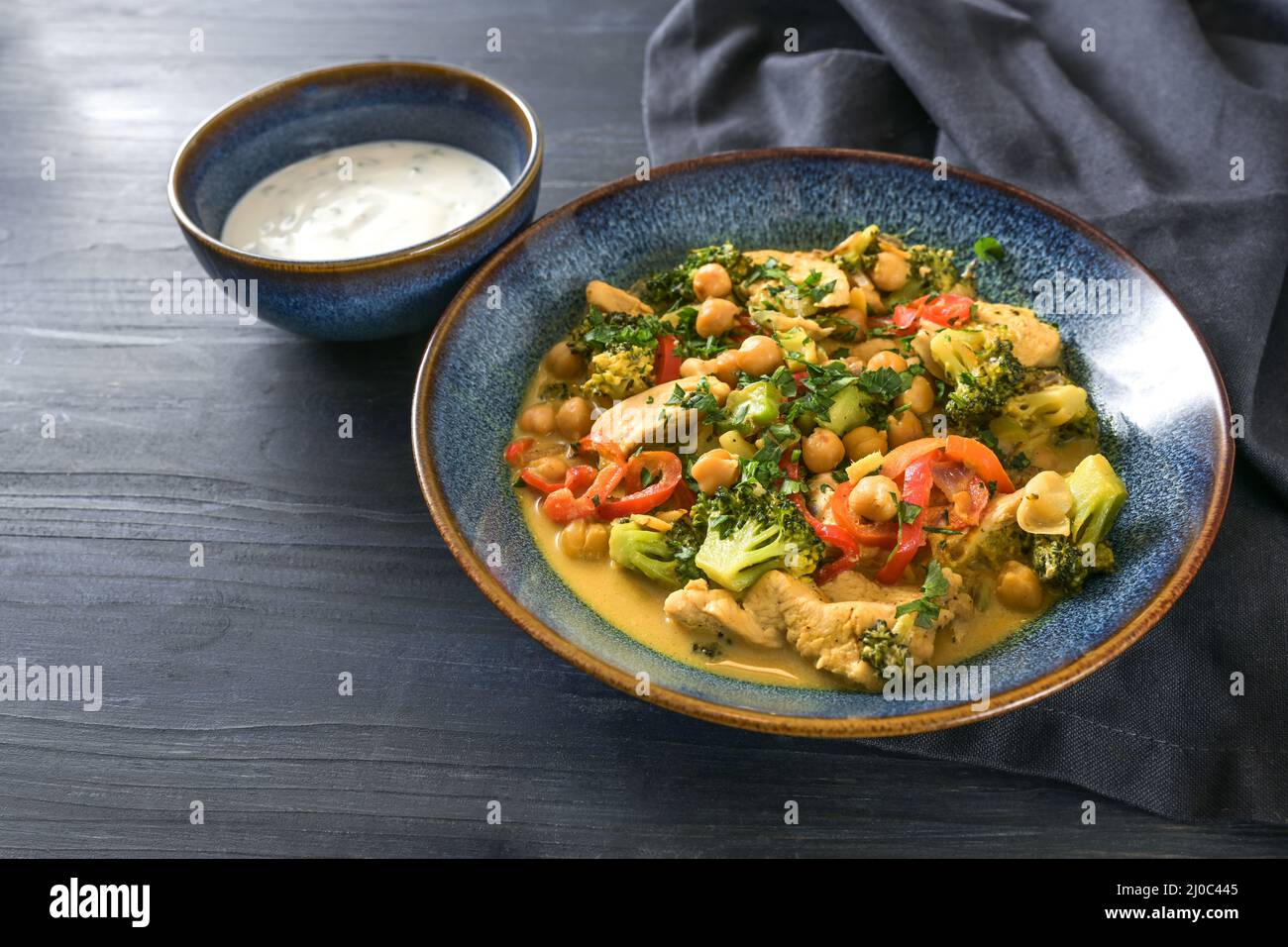 In einer separaten Schüssel auf einem dunkelblauen Holztisch, mit Kopierraum, ausgewähltem Fokus, enger Tiefe, eine Mahlzeit aus Kichererbsen, Gemüse und Huhn mit Joghurt Stockfoto