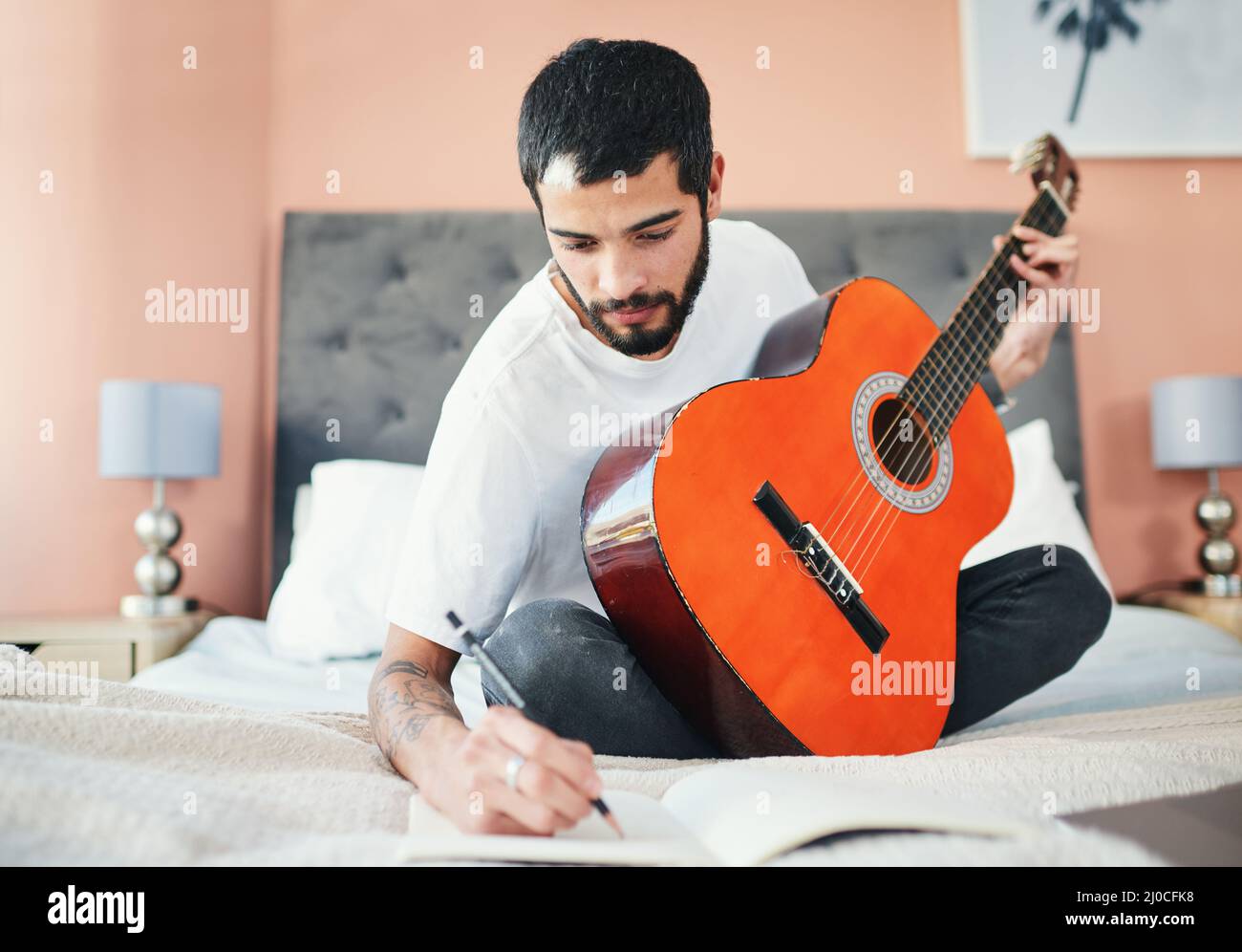 Ich bin mir sicher, dass dies ein Hit werden wird. Aufnahme eines Mannes, der in seinem Notizbuch schreibt, während er zu Hause Gitarre spielt. Stockfoto