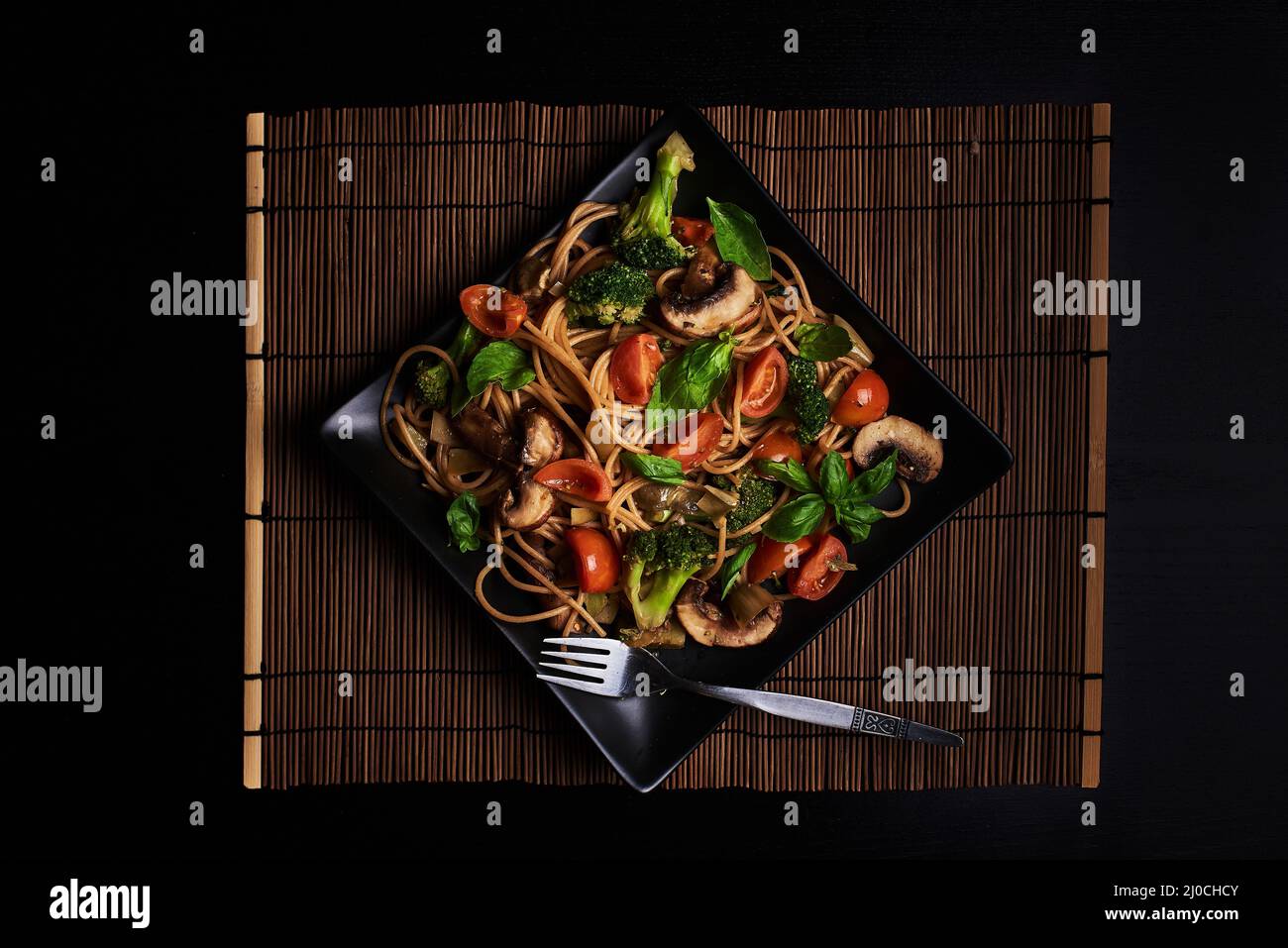 Quadratischer Teller mit italienischer Spaghettis mit Tomaten, Brokkoli, Pilzen und Basilikumblättern. Draufsicht auf dunklem Hintergrund. Vegane gesunde Pasta-Mahlzeit Stockfoto