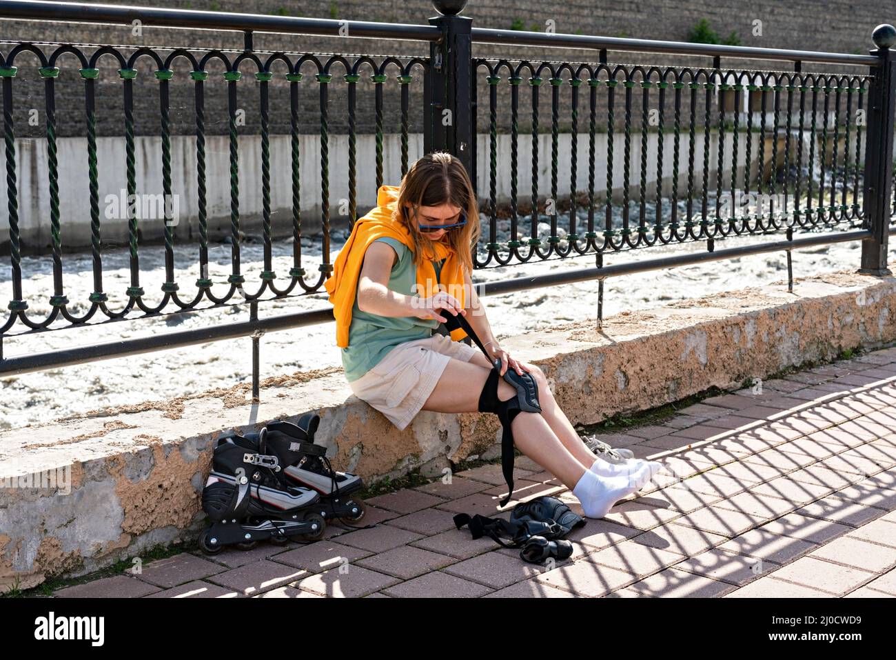 Junge Frau im gelben Hoodie, die Schutzausrüstung anlegt, bevor sie am Ufer Rollschuhlaufen kann aktiver Lebensstil, Outdoor-Aktivitäten, Rollschuhlaufen Stockfoto