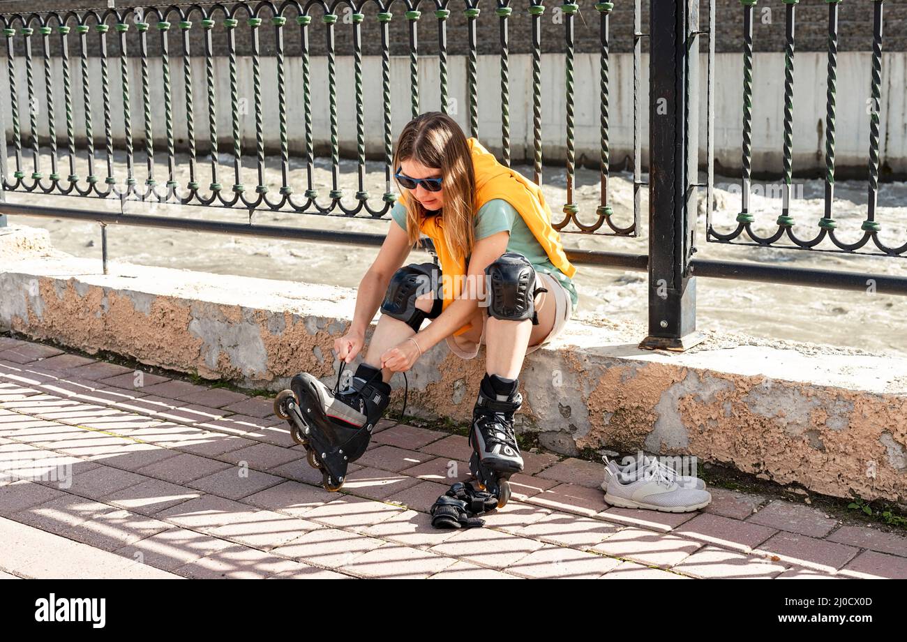 Junge Frau im gelben Hoodie, die Schutzausrüstung anlegt, bevor sie am Ufer Rollschuhlaufen kann aktiver Lebensstil, Outdoor-Aktivitäten, Rollschuhlaufen Stockfoto