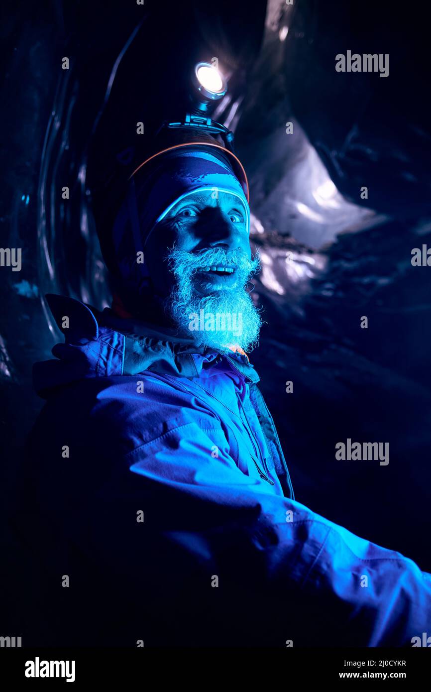 Portrait des alten Alpinisten mit grauem Bart im Helm mit glühender Stirnlampe lächelnd in tiefdunkler Gletscherhöhle mit blauen Lichtern erkunden Winterberg g Stockfoto