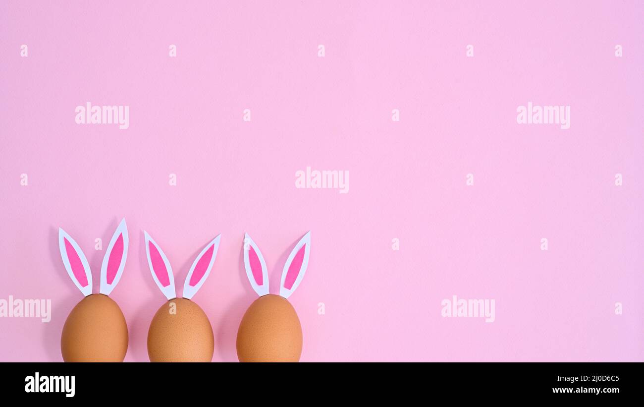 Drei natürliche Eier mit Kaninchenohren schauen auf pastellrosa Hintergrund mit Kopierraum. Ostern flach lag minimal Stockfoto
