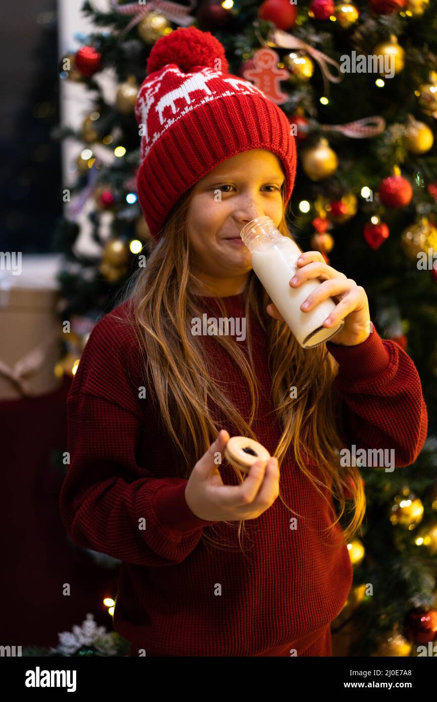 Kind in roten Weihnachtskleidung und Winterpullover trinkt Milch aus einer Glasflasche wie Santa. .Horizontales Foto für Weihnachten, Neujahr, Weihnachten. Stockfoto