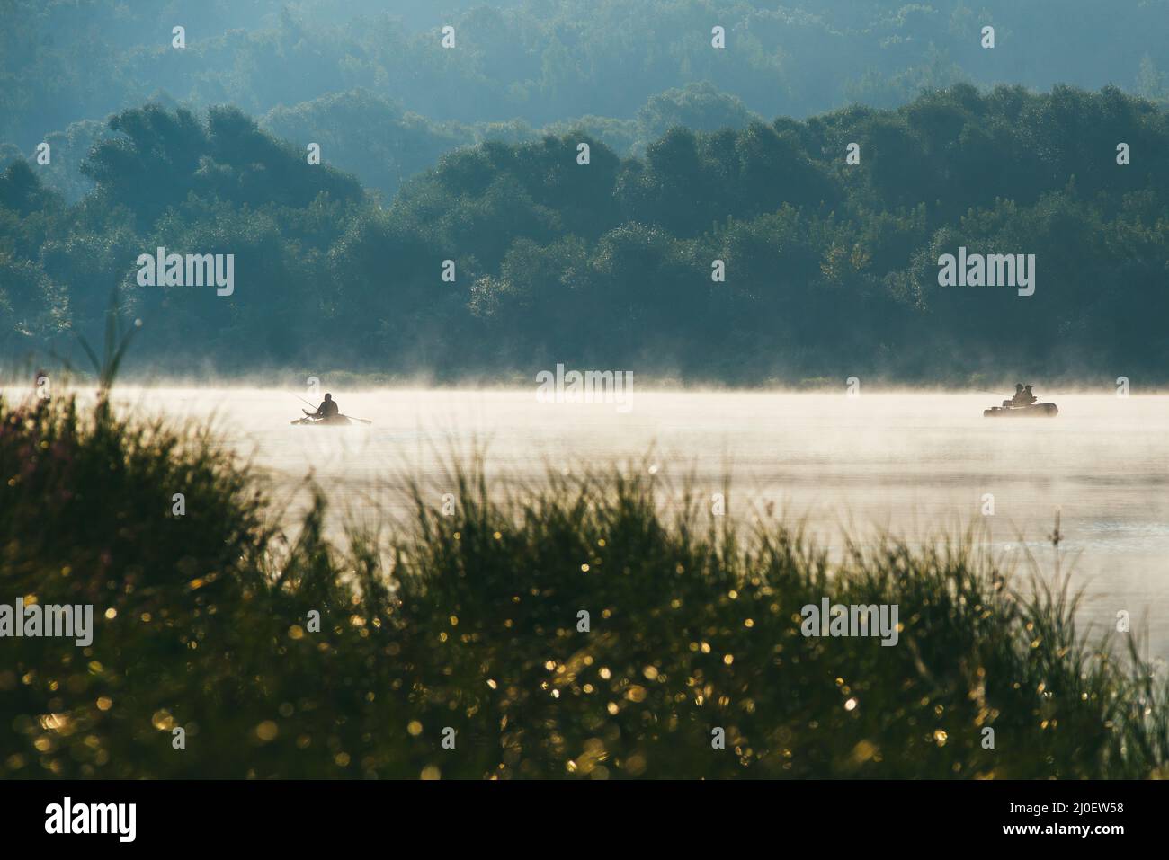 Fischermänner fischen auf einem Boot. Frühmorgens Frühlingslandschaft mit Fluss im Vordergrund und einem grünen Ufer mit Büschen und Bäumen Stockfoto