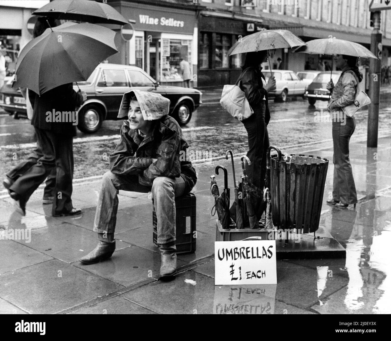 Ein Bild aus einer Serie humorvoller Neuheitenbilder, aufgenommen vom Sunday People-Fotografen Dennis Hutchinson. Ein Straßenhändler verkauft Regenschirme bei Regen. Er benutzte eine Zeitung anstelle eines Regenschirms für sich selbst. Circa: 1978 Stockfoto