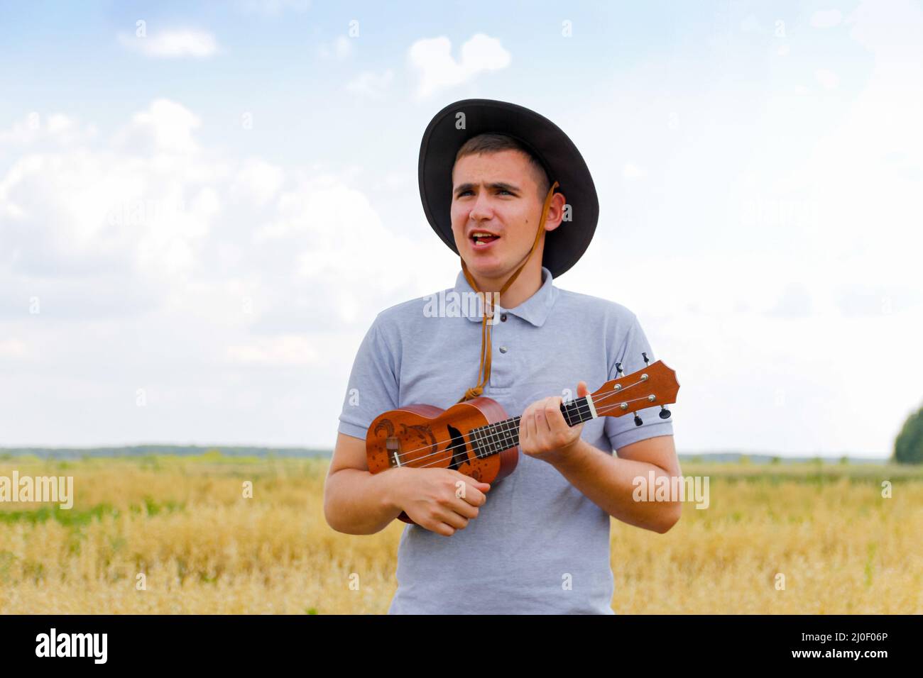 Fröhlicher Mann. Junger schöner Kaukasischer, der draußen Ukulele spielt. Schöner Hipster Kerl spielt Ukulele Gitarre auf Natur Hintergrund. Männchen in einem Cowboy Stockfoto