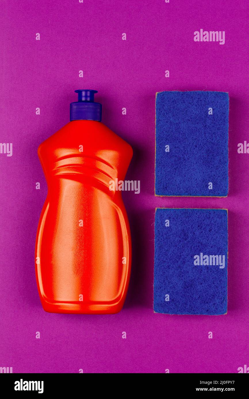 Orangefarbene Plastikflasche mit Spülmittel und blauem Schwamm auf violettem Hintergrund, Nahaufnahme von oben Stockfoto