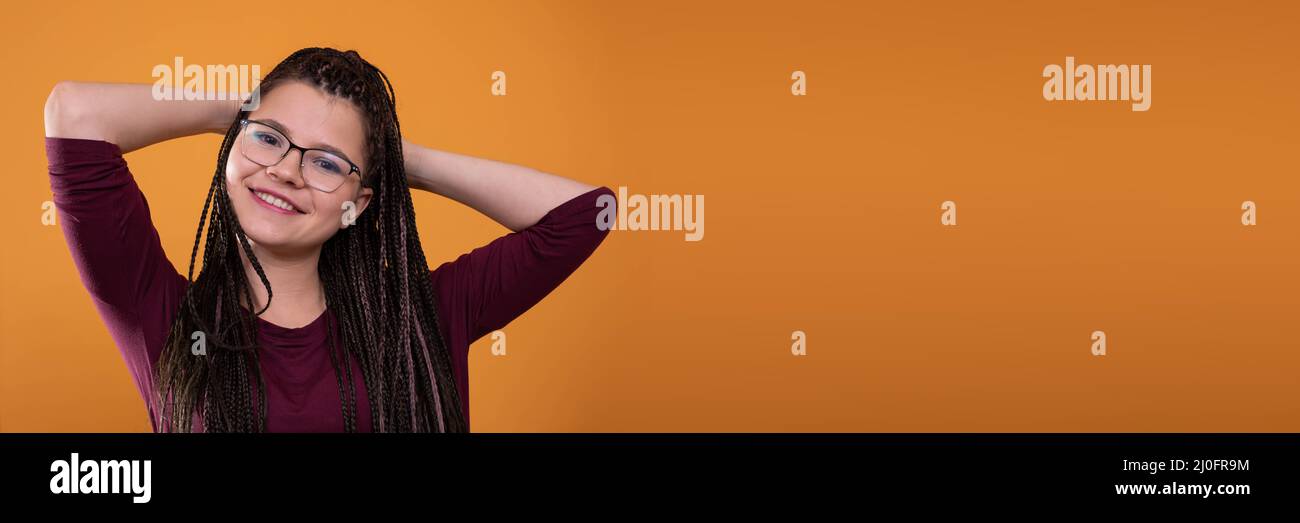 In einer Panoramaaufnahme lächelt ein junges Mädchen mit einer Brille und den Händen auf dem Kopf aufrichtig. Orange verdünnter Hintergrund. Charakter Stockfoto