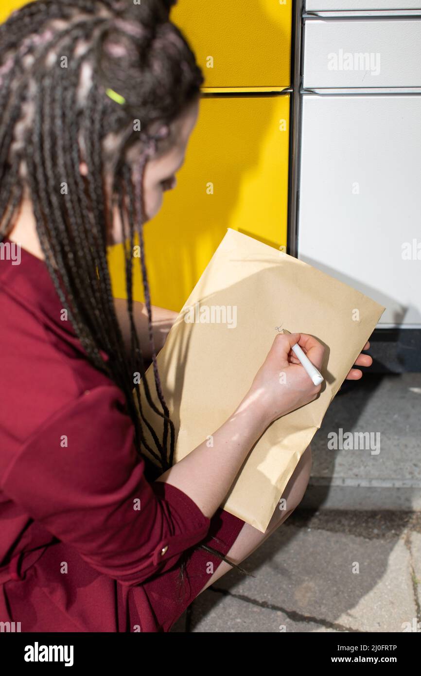 Ein junges Mädchen spricht den Brief an, während es neben dem Paketfach steht. Verwenden Sie einen Kugelschreiber, um auf den grauen Buchstaben zu schreiben Stockfoto