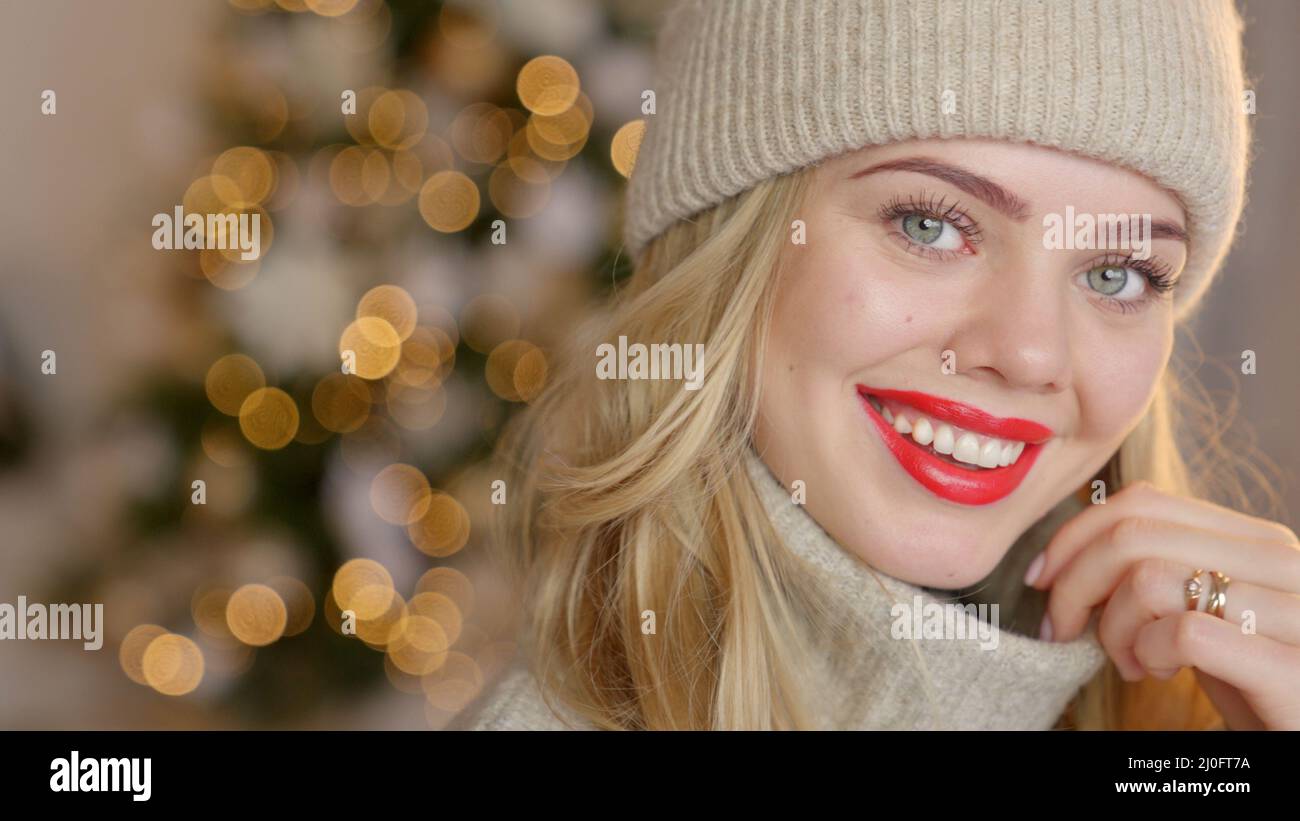 Lächelnde blonde Frau mit rotem Lippenstift, in einer warmen Wolljacke gekleidet und Hut posiert in ihrem Haus Stockfoto