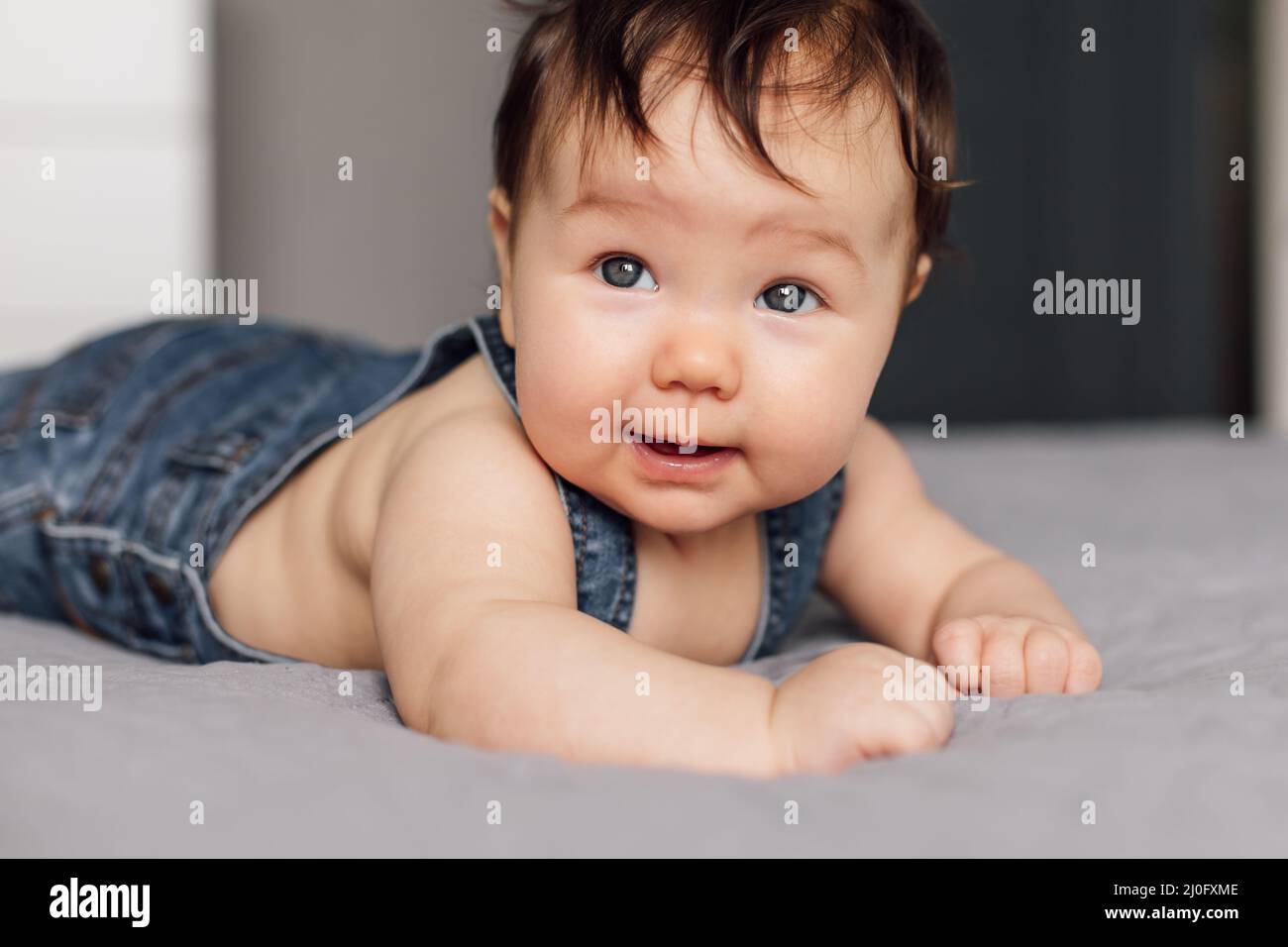 Porträt eines Babys in Denim-Strampler, der zu Hause auf einer grauen Decke liegt. Das blauäugige Kleinkind, das im Schlafzimmer auf dem Bett kriecht, erkundet die Welt Stockfoto