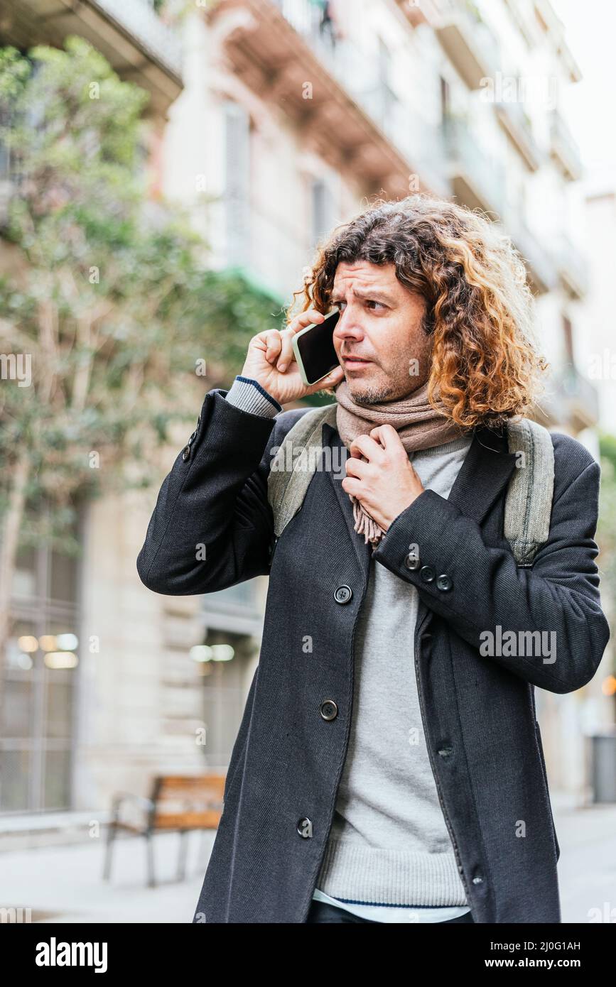 Mann mittleren Alters in eleganter Freizeitkleidung mit lockigem Haar, das den Schal berührt und wegschaut. Er geht auf der Stadtstraße und nimmt tagsüber einen Anruf an Stockfoto