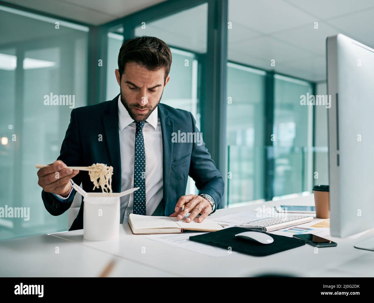 Kann sich nicht auf eine Sache auf einmal konzentrieren. Aufnahme eines fokussierten jungen Geschäftsmannes, der Nudeln aß, während er in einem Notizbuch im Büro schrieb. Stockfoto