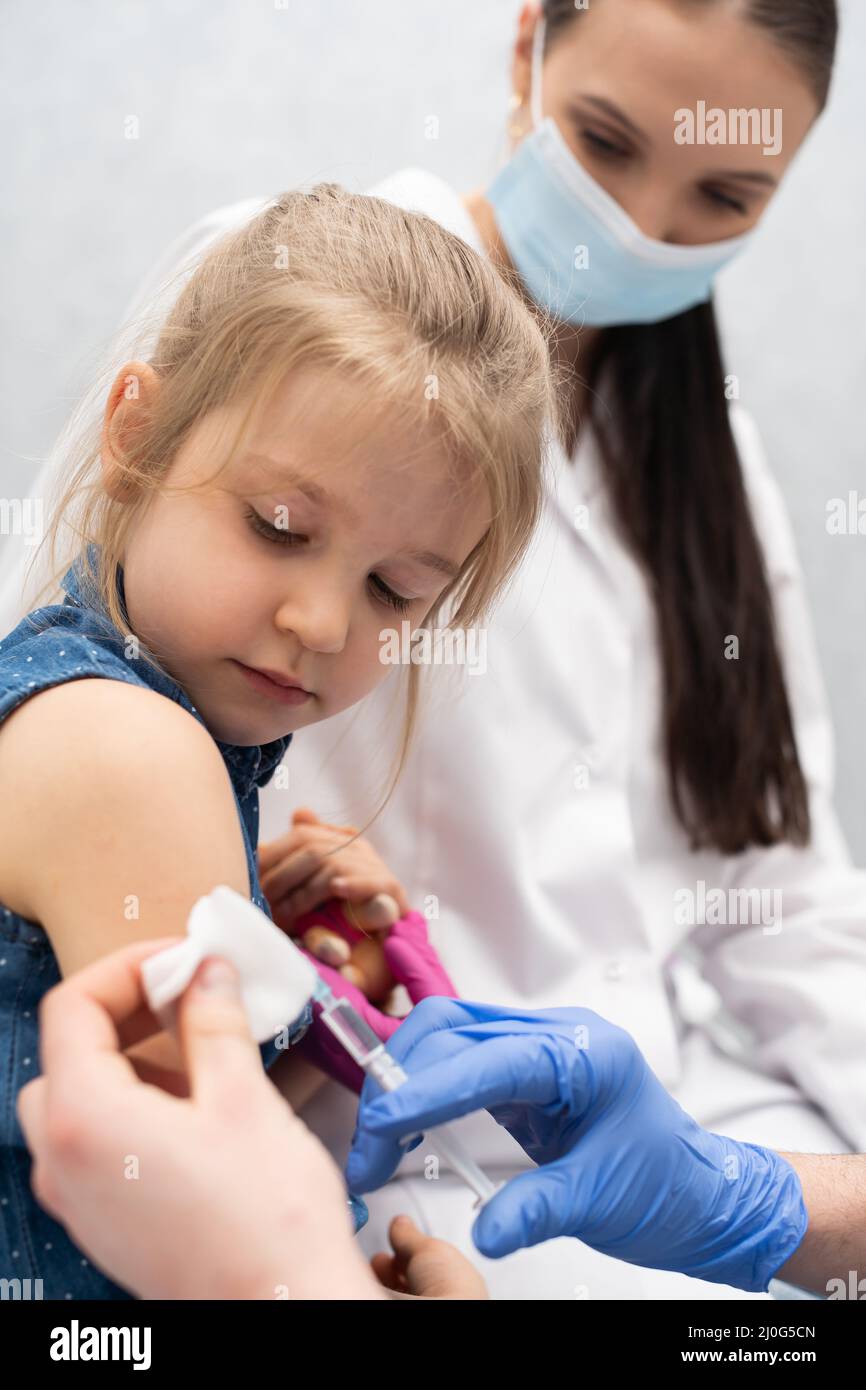 Der Arzt macht sich bereit, ein kleines Mädchen zu impfen. Eine junge Krankenschwester sitzt neben dem Kind, damit sich das Kind sicher fühlt. Preven Stockfoto