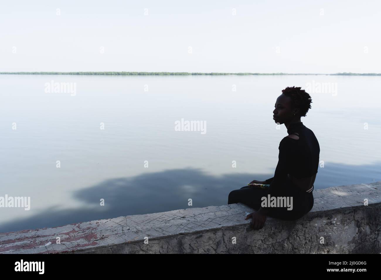 Silhouette eines einsamen jungen afrikanischen Teenagers, das auf einer Wand am Fluss sitzt und auf das andere Ufer blickt; Symbole der Einsamkeit, der Sehnsucht, des Nostalgis Stockfoto