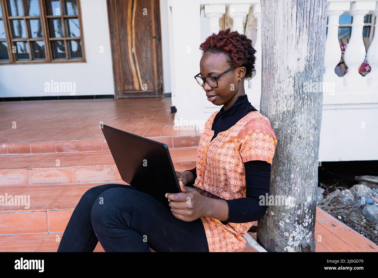 Intelligentes, junges afrikanisches Mädchen mit großen Gläsern, das auf den Stufen der Veranda ihres Hauses sitzt, während der COVID-Sperre im Fernunterricht Stockfoto