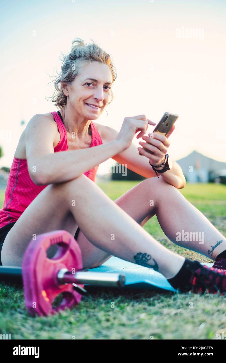 Echte junge reife kaukasische Frau in einer Sportkleidung im Park, die ihr Handy an einem sonnigen Nachmittag sucht. Stockfoto