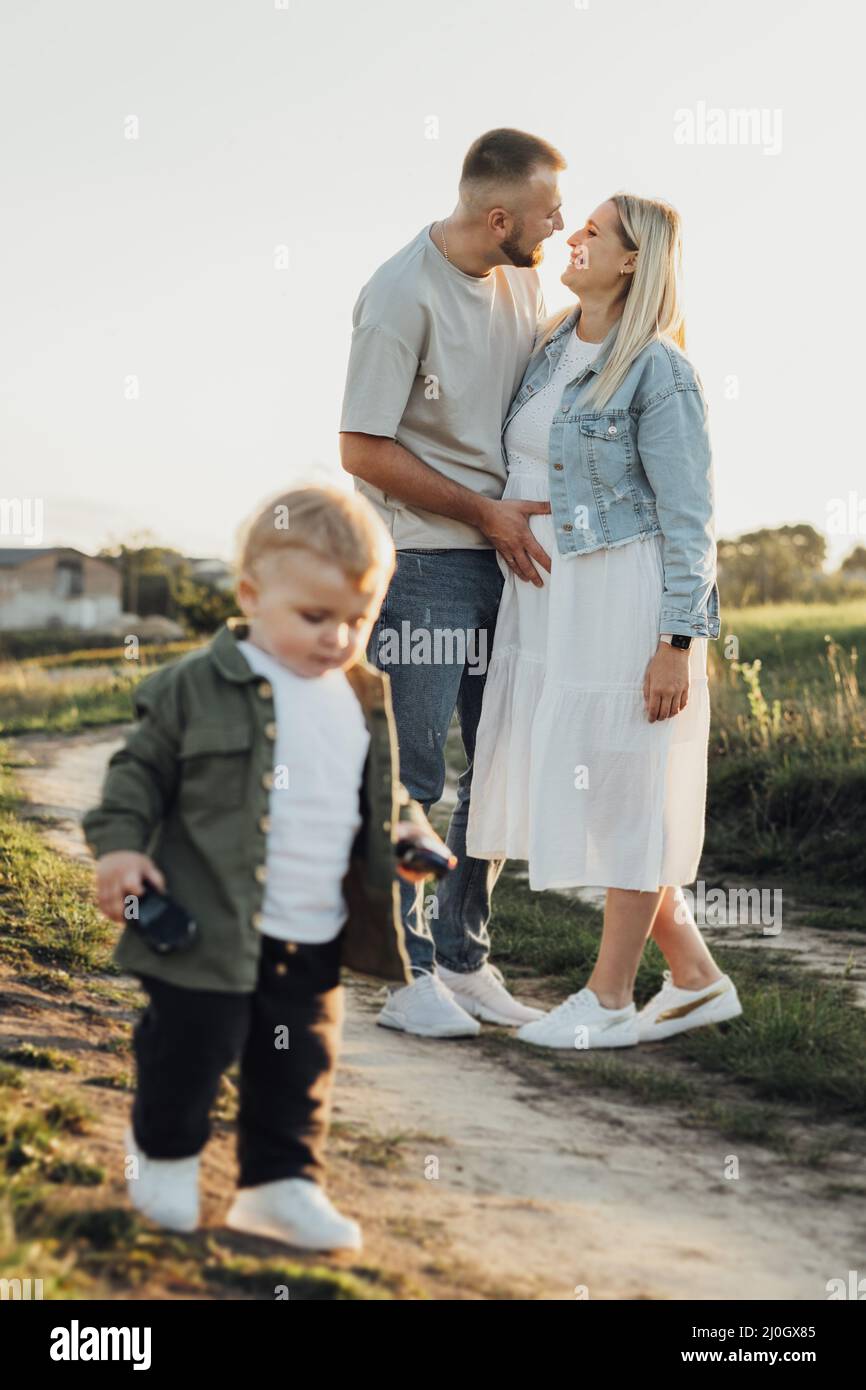 Glückliche junge Familie im Freien, Vater hält seine Schwangere durch Bauch, während ihr Kleinkind Sohn um sie herum spielt Stockfoto