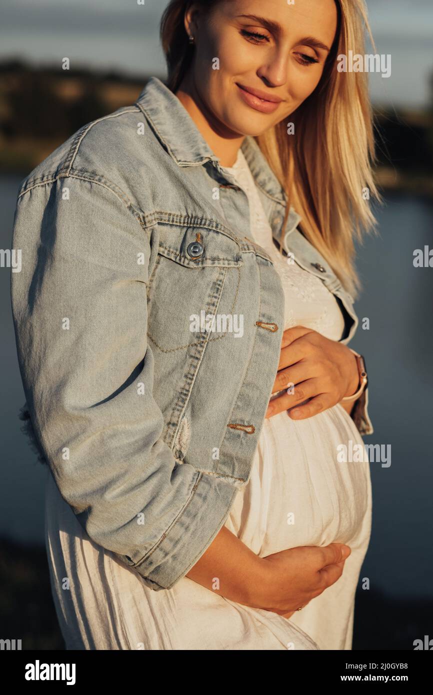 Nahaufnahme Porträt einer Schwangeren jungen kaukasischen Frau, die ihren Bauch bei Sonnenuntergang im Freien hält Stockfoto