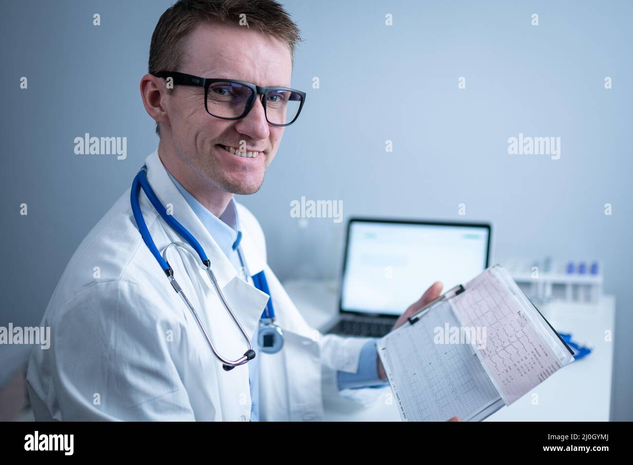 Junger, gutaussehender Arzt mit Brille und weißem Laborkittel untersucht die Ergebnisse der elektrokardiographischen Untersuchung des Patientenherzens in Stockfoto