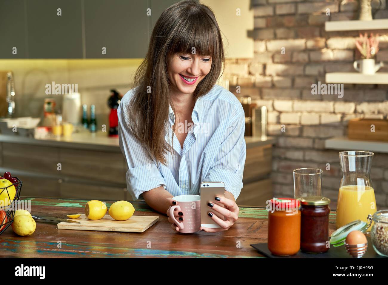 Glückliche junge attraktive weiße Frau im Männerhemd, die am Morgen mit dem Handy in der Küche zu Hause frühstückte und lächelte. Stockfoto