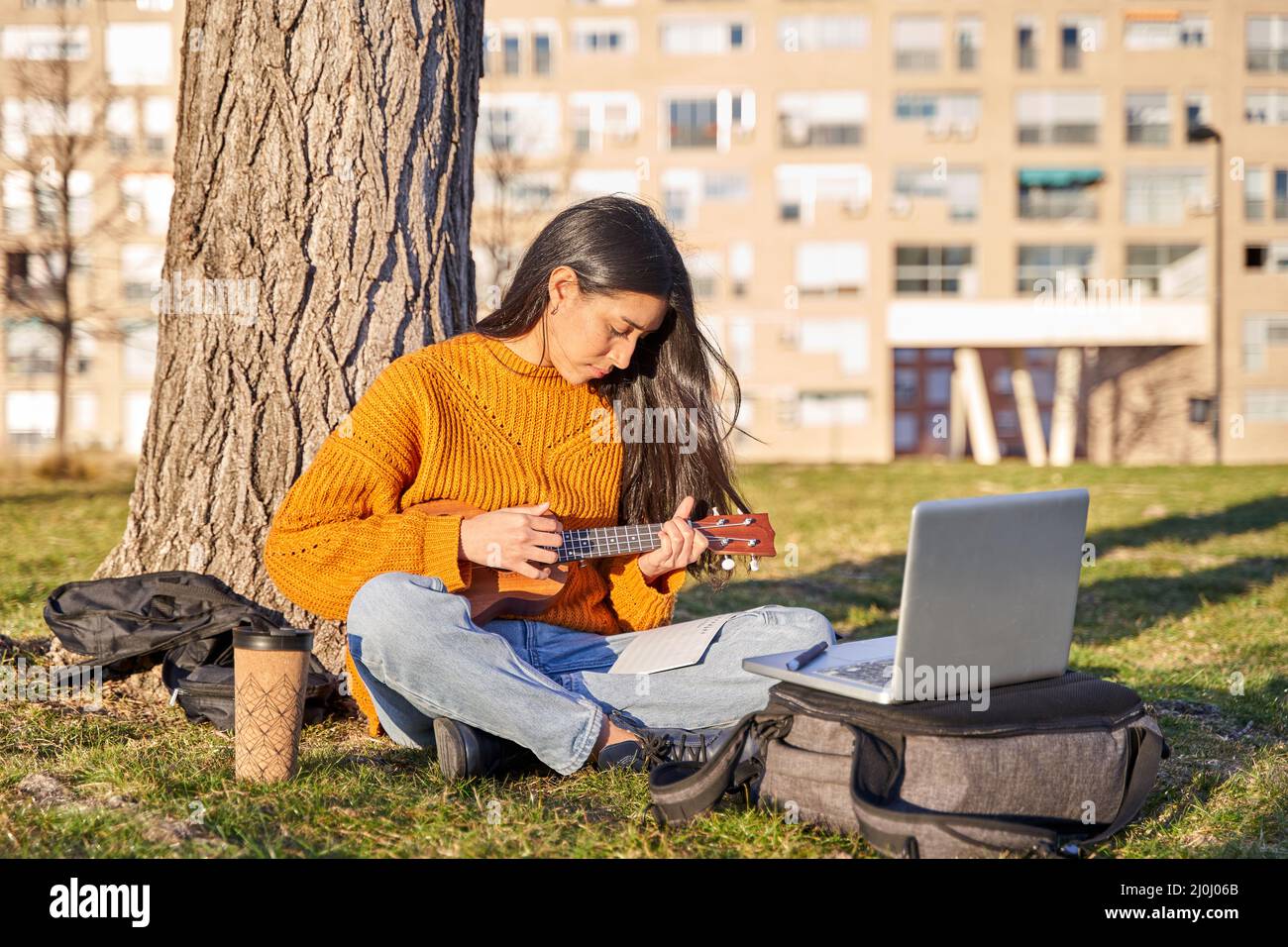 Junge Latina mit langen Haaren spielt die Ukulele unter einem Baum. Sie hat einen Laptop, um den Leccion zu beobachten. Musikkonzept und lernen Autodidakt Stockfoto