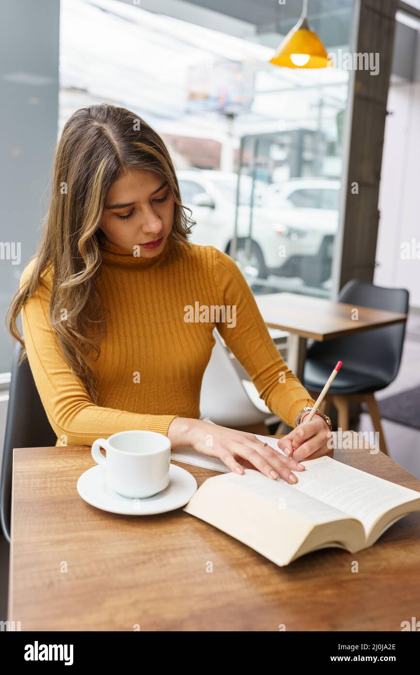 Lernen und arbeiten beim Schreiben eines Buches mit einem Bleistift eine schöne junge latinerin mit langen blonden Haaren sitzt in einer Cafeteria mit einer Tasse Stockfoto