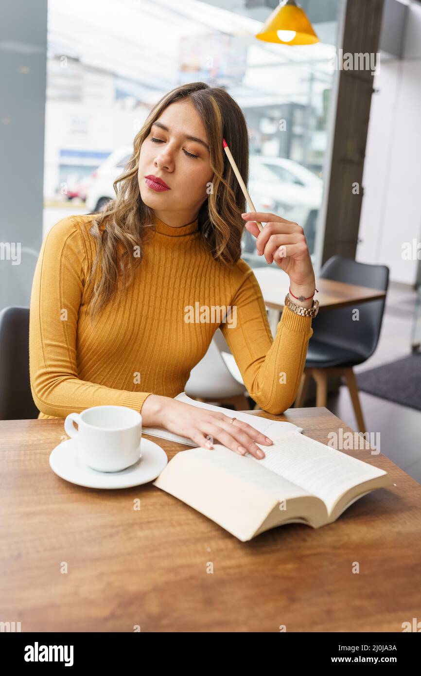 Mit einem Bleistift auf dem Kopf denkend eine junge lateinerin mit langen blonden Haaren sitzt in einer Cafeteria mit einer Tasse Kaffee, studentischer Lebensstil Stockfoto