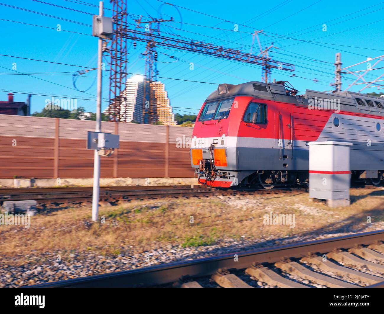 Die Vorderseite eines Zuges, der an einem sonnigen Tag vor der Kulisse eines Zauns und eines Gebäudes entlang der Schienen rast Stockfoto