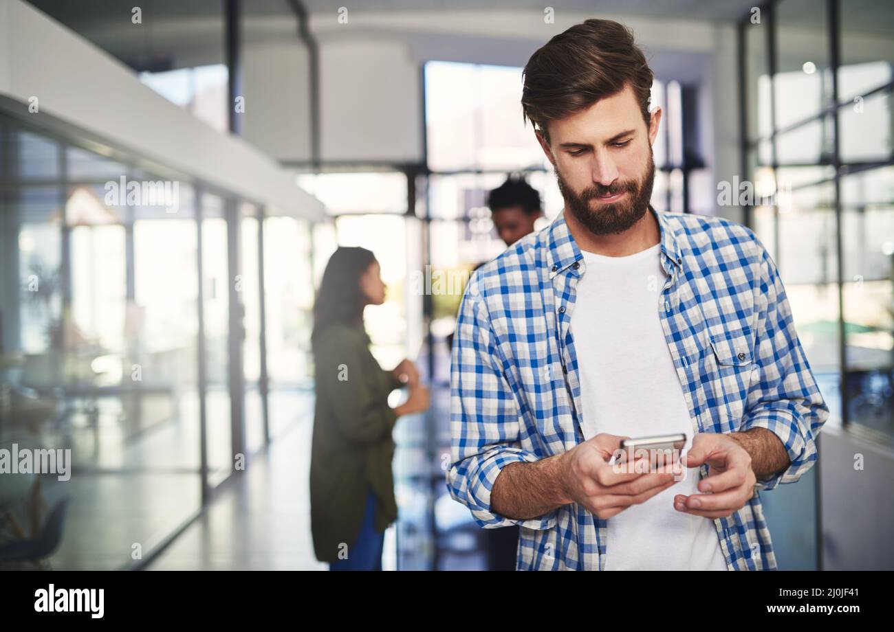 SMS an einen Client mit den neuesten Updates senden. Ausgeschnittene Aufnahme eines jungen Designers, der ein Mobiltelefon in einem Büro mit Kollegen im Hintergrund verwendet. Stockfoto