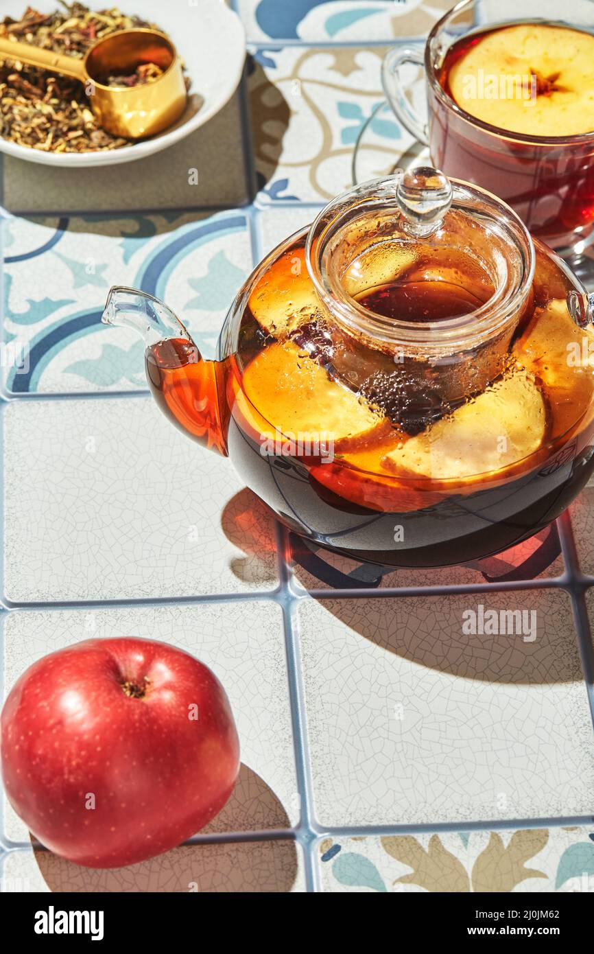 Früchtetee mit Äpfeln und Thymian in Glas Teekane und Tasse auf Tisch aus farbigen Fliesen Stockfoto