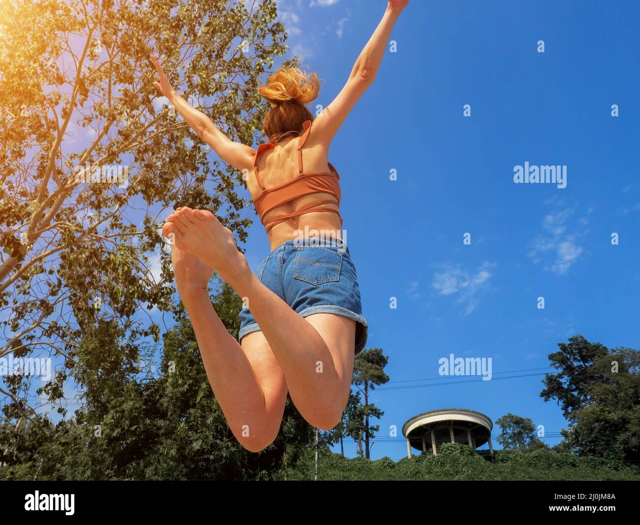 Ein Mädchen in einem Sprung mit gespreizten Armen und zusammengerollten Beinen vor einem Hintergrund von blauem Himmel und grünem Park Stockfoto