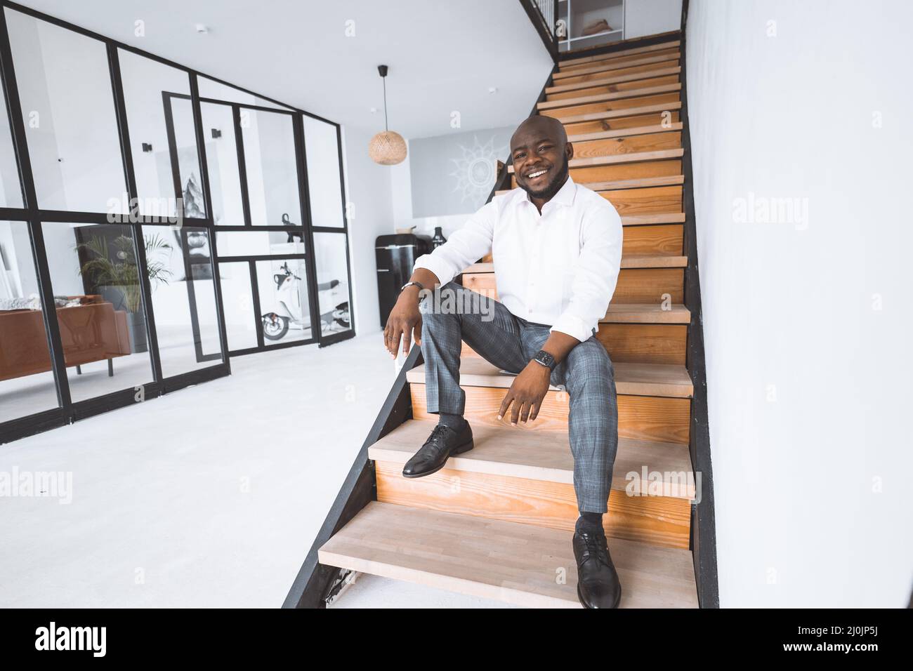 Happy Young Erfolgreicher Geschäftsmann In Seinem Erworbenen Haus. Afro American sitzt auf einer Treppe in Hall in Apartments und Smiles. Concep Stockfoto
