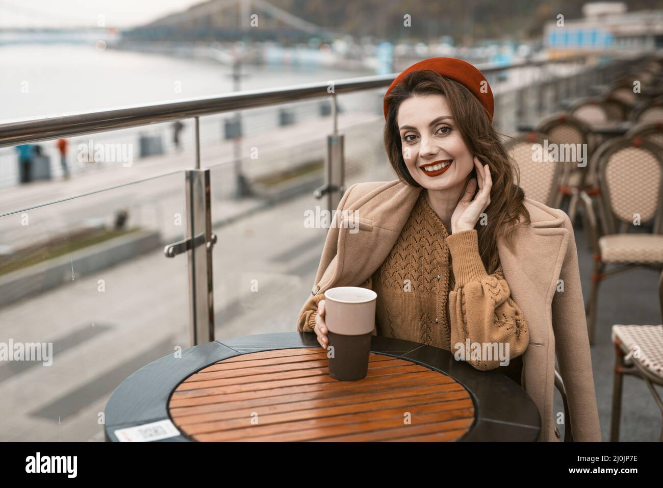 Glückliches junges Mädchen, das draußen im Straßenrestaurant oder auf der Terrasse sitzt und lächelnd einen Kaffeebecher hält. Porträt eines stilvollen Jos Stockfoto