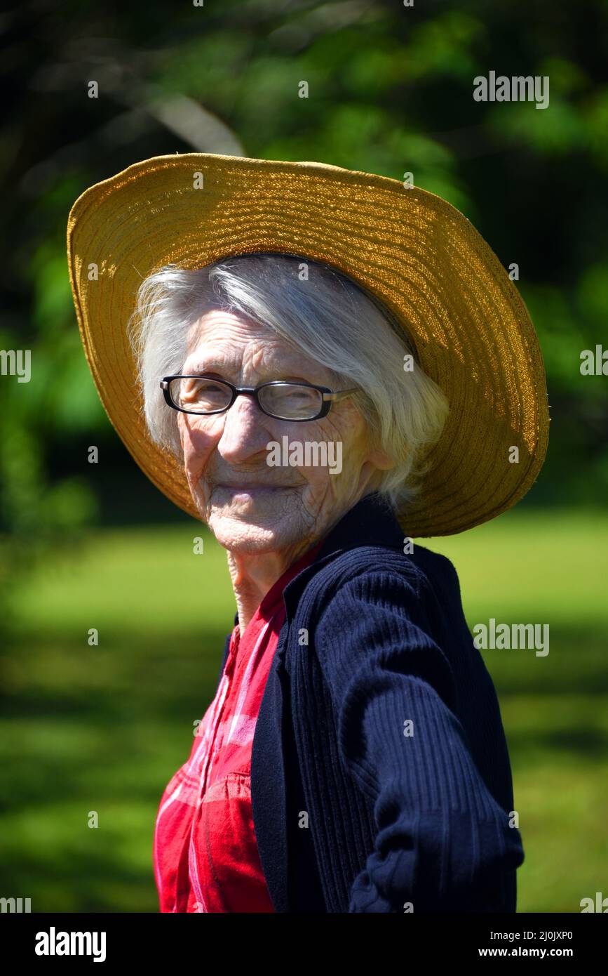 Eine hundertjährige Landfrau steht im Freien in ihrem roten karierten Hemd und Strohhut. Die Sonne scheint auf ihrem weißen Haar. Stockfoto